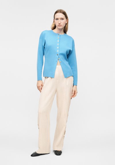 Γυναίκα με ίσια ξανθά μαλλιά φοράει μπλε ζακέτα με κουμπιά και εκρού παντελόνι φαρδιάς γραμμής, στέκεται με ουδέτερη έκφραση σε λευκό φόντο.