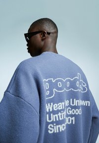 Persoon met zwarte zonnebril en een blauwe trui met witte tekst "goods Wear Untwun Until Good Since 001" op de rug.