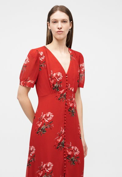 Pruunide sirgete juustega naine, kes kannab punast lillelist kleiti, millel on pufflõhiseid varrukaid ja ees kangaga kaetud nööbid, ühevärvilisel taustal.