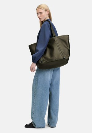 Olivgrüne Tragetasche aus glattem Stoff mit strukturiertem Design und doppelten Griffen. Das Modell trägt weite blaue Jeans und ein marineblaues Oberteil.