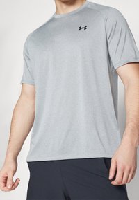 Mężczyzna w jasnoszarej koszulce sportowej z krótkim rękawem z czarnym logo Under Armour oraz czarnych szortach, z ramionami luźno opadającymi wzdłuż ciała.