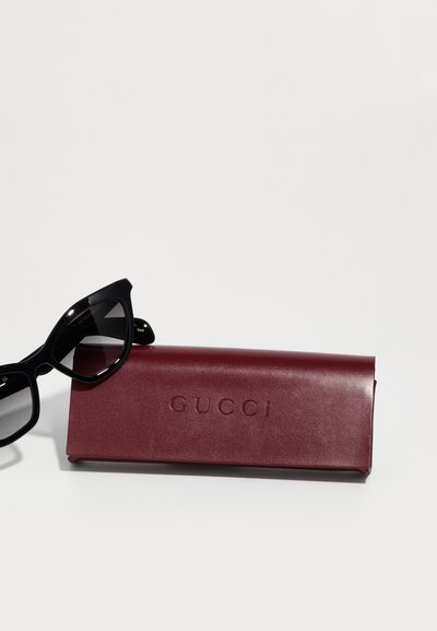 Occhiali da sole neri a forma di occhio di gatto accanto a un astuccio in pelle marrone bordeaux con logo "GUCCI" impressa, texture liscia e design elegante.
