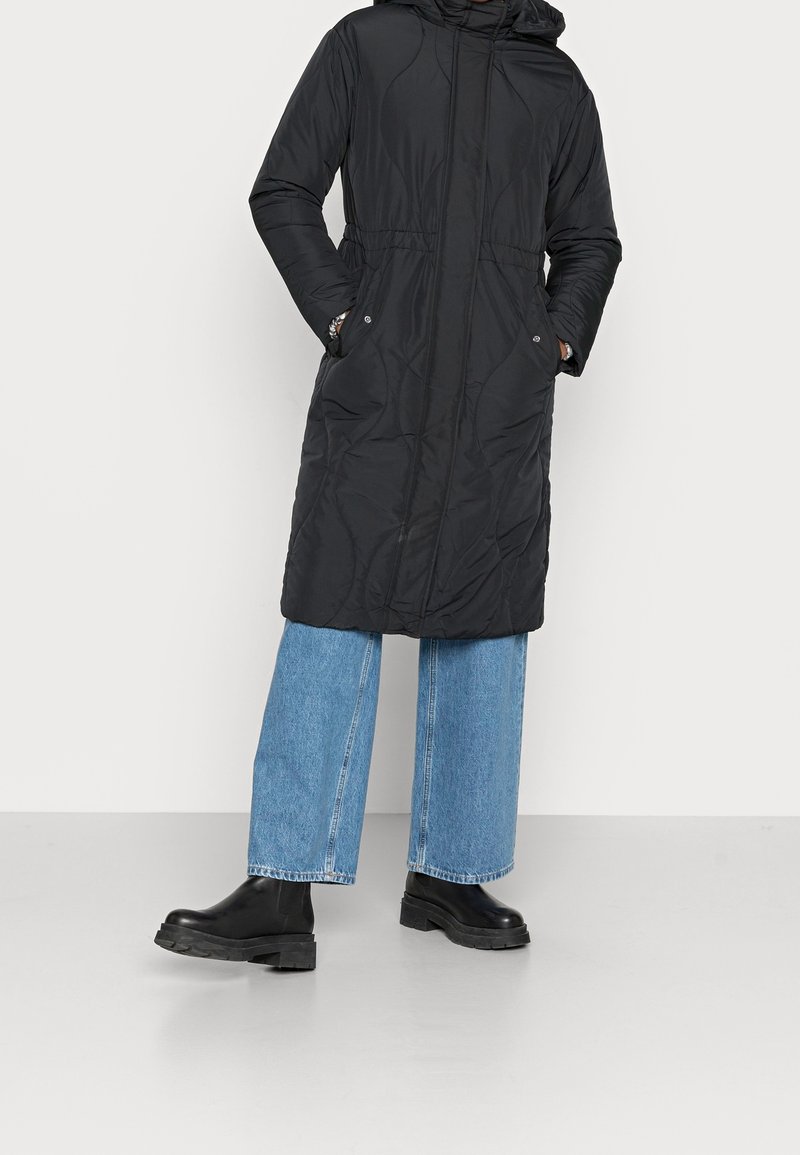 Dlouhý černý péřový kabát s kapucí, zúžený v pase, s bočními kapsami a prošívaným vzorem. Kombinován se širokými modrými džínami a černými botami.
