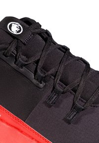 Scarpa sportiva nera con accenti rossi, caratterizzata da una tomaia testurizzata, un sistema di allacciatura tradizionale e un logo circolare sulla linguetta.