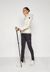 Svetlokrémová bunda na zips s kapucňou a čiernymi detailmi, skombinovaná s čiernymi nohavicami úzkeho strihu a béžovými teniskami. Drží trekingové palice.