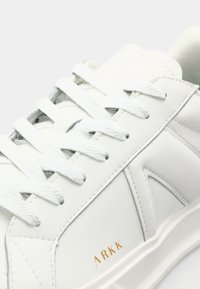 White leather sneaker with flat white laces, textured panels, and gold "ARKK" branding on the side. Smooth surface and minimalistic design.