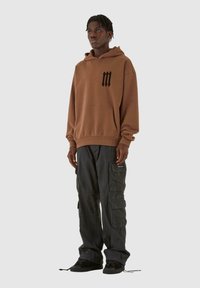 Felpa in cotone marrone con logo nero sul petto, abbinata a pantaloni cargo neri con molte tasche e sneakers nere.