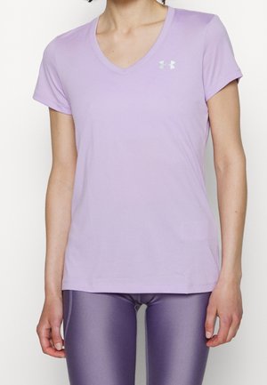 Kobieta nosząca jasnofioletowy sportowy T-shirt z dekoltem w serek i krótkim rękawem oraz błyszczące ciemnofioletowe legginsy, stojąca z luźno opuszczonymi rękami przy bokach.