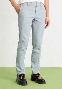 Lichtgrijze tailored broek met een slim fit, platte voorkant en riemlussen, gecombineerd met zwart en bruin gespikkelnde schoenen, op een groene vloer.