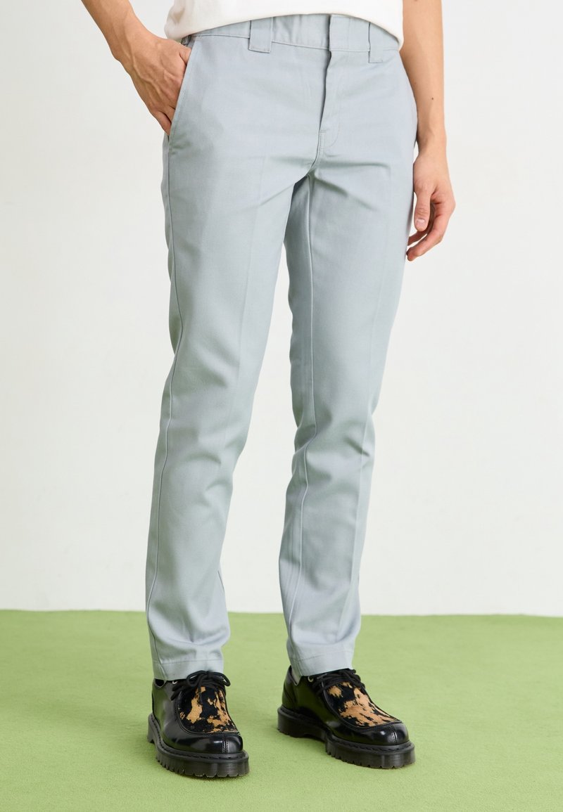 Lichtgrijze tailored broek met een slim fit, platte voorkant en riemlussen, gecombineerd met zwart en bruin gespikkelnde schoenen, op een groene vloer.