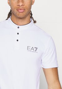 Biele polo tričko s krátkym rukávom a zapínacím golierom, ktoré má tri čierne gombíky a čierne výšivkové logo na ľavej strane.