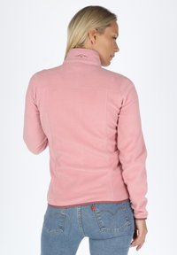 Giacca in pile rosa con colletto alto, dettagli cuciti e logo sul retro, abbinata a jeans blu. Texture liscia.