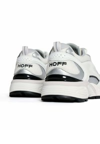 Witte en zilveren sport sneakers hebben een gestructureerde bovenkant, zwarte accenten en het "Hoff" logo op de hiel, met een dikke rubberen zool.