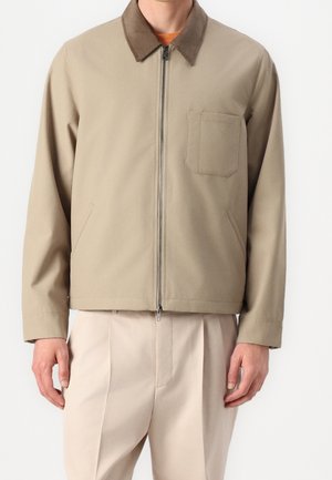 Beige lynlåsjakke med brun krave og brystlomme, båret over en orange skjorte og kombineret med lysebeige bukser.