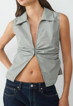 Gestreifte ärmellose Bluse in Olivgrün und Weiß, mit tiefem V-Ausschnitt, geknoteter Front und abgerundetem Saum. Kombiniert mit dunkelblauen Jeans.