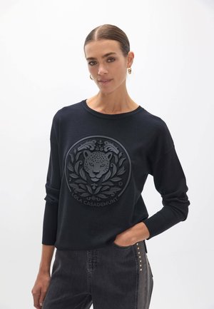 Mujer con cabello oscuro en un moño bajo lleva un suéter negro con leopardo en relieve y hojas decorativas, de pie con una mano en el bolsillo.