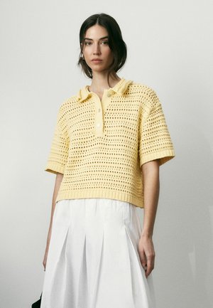 Žena nosi voľnú žltú háčkovanú polokošeľu a bielu plisovanú sukňu na jednoduchom pozadí.