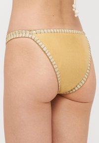 Żółty dół bikini z błyszczącym złotym wykończeniem, o gładkiej fakturze i niskim kroju. Widok z tyłu podkreśla krój i detale.