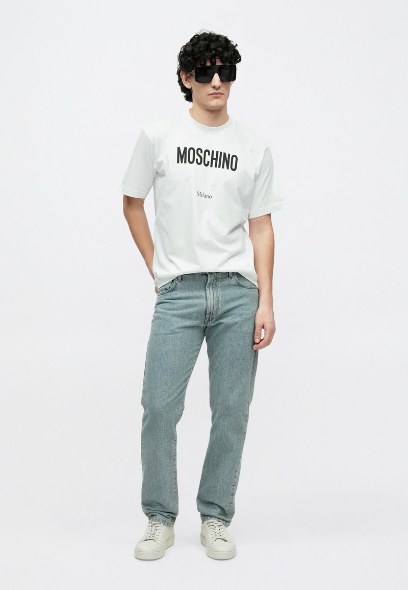 Hvid t-shirt med "MOSCHINO Milano"-print, blå denimjeans og beige sneakers. Model iført store sorte solbriller med krøllet hår.