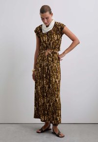 Femme portant une robe deux pièces à motifs bruns avec découpes à la taille, un foulard blanc au cou, un bracelet beige et des sandales noires cloutées, se tenant à l'intérieur.