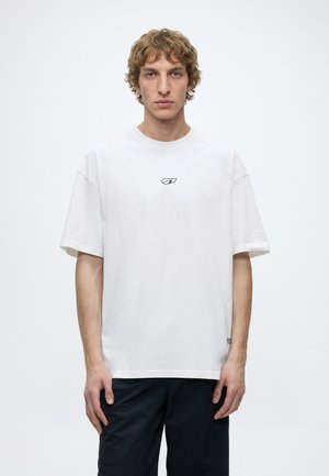 Junger Mann mit welligem blondem Haar, der ein weißes oversized T-Shirt mit kleinem Logo und schwarze Hose trägt, vor einem schlichten weißen Hintergrund steht.