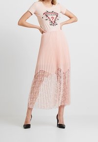 T-shirt rose avec un graphique "GUESS?", associé à une jupe plissée en couches rose et en dentelle. Des escarpins noirs complètent la tenue.
