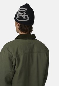 Veste en velours côtelé verte avec un bonnet en tricot noir arborant un motif géométrique gris. La veste possède un logo brodé dans le dos.