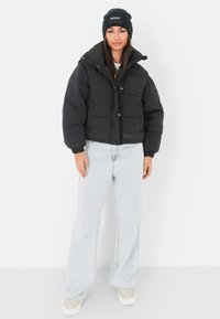 Czarna kurtka puchowa o skróconym kroju z wyraźnymi przeszyciami, zestawiona z jasnoniebieskimi jeansami z wysokim stanem i czarną czapką beanie.