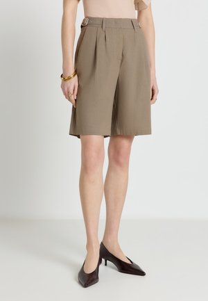 Short beige taille haute ajusté longueur genou avec boucles latérales, porté avec des escarpins marron foncé à bout pointu et des bijoux dorés au poignet et à la main.
