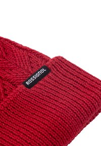 Rode gebreide beanie met een gestructureerd patroon en een zwart label met de tekst "ROSSIGNOL." Heeft een strakke pasvorm en een gelijkmatige, zachte textuur.