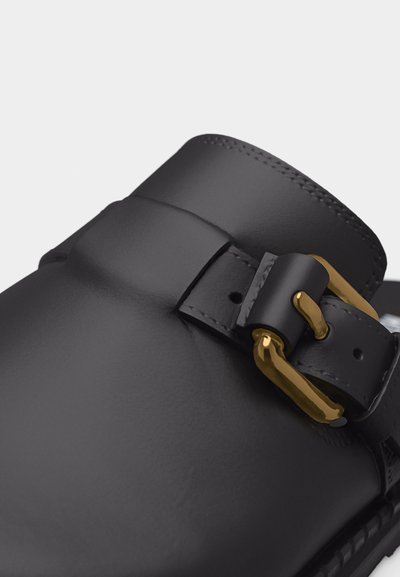 Gros plan sur une chaussure en cuir noir avec une boucle en métal doré et un détail de sangle sur la partie supérieure de la chaussure.