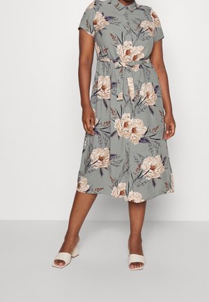 Femme portant une robe florale grise longueur genou, à manches courtes, boutonnée devant et ceinturée à la taille, associée à des sandales blanches à talon bloc et à bout ouvert.