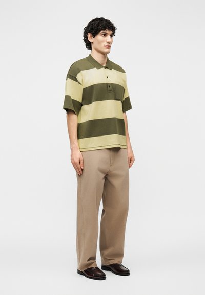 Άνδρας που στέκεται φορώντας υπερμεγέθη ριγέ πουλόβερ πόλο σε ελιά πράσινο και μπεζ, χαλαρά μπεζ παντελόνια και σκούρα καφέ παπούτσια, μπροστά σε απλό φόντο.