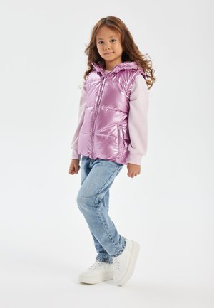 Gilet puffo rosa, lucido, con cappuccio, chiusura con zip e sezioni gonfie, indossato sopra una maglietta a maniche lunghe rosa chiaro e jeans blu. Sneakers bianche.