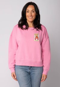 Vrouw met donker haar draagt een roze sweatshirt met een kleurrijke koffiethema-patch, gecombineerd met lichtblauwe jeans, glimlachend tegen een neutrale achtergrond.