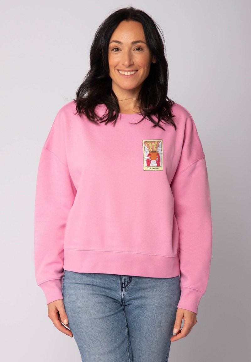 Vrouw met donker haar draagt een roze sweatshirt met een kleurrijke koffiethema-patch, gecombineerd met lichtblauwe jeans, glimlachend tegen een neutrale achtergrond.