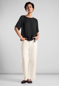 Schwarzes T-Shirt mit kurzen Ärmeln, rundem Halsausschnitt und Kordelzugdetails, kombiniert mit weißen, weit geschnittenen Hosen und schwarzen flachen Schuhen.