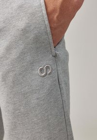 Gray sweatpants featuring a side pocket and an embroidered logo in a lighter shade. Soft, smooth texture with a relaxed fit.