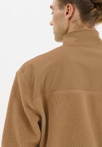 Veste en polaire marron avec une moitié inférieure en shearling texturé et une moitié supérieure lisse et légère, dotée d'un col haut.