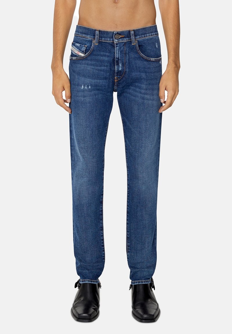 Homme portant un jean denim bleu avec des détails légèrement usés et des chaussures en cuir noir, debout devant un fond clair uni.