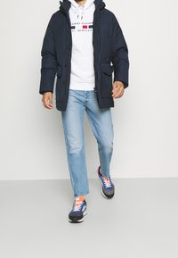 Marinblå vadderad jacka med framknappar, två stora fickor, i kombination med en vit hoodie och ljusblå jeans. Färgglada sneakers med kontrasterande snören.