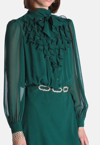 Femme portant un chemisier long à manches longues en voile vert foncé avec des volants et un nœud, associé à une ceinture chaîne scintillante et un bracelet assorti.