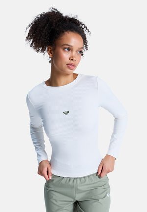 Haut blanc à manches longues côtelé avec un col rond et un logo cœur vert sur la poitrine, associé à un pantalon ample vert clair.