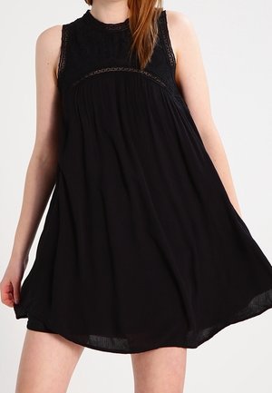 Mujer que lleva un vestido negro sin mangas con detalles de encaje en la parte superior, sosteniendo la falda fluida ligeramente hacia fuera.