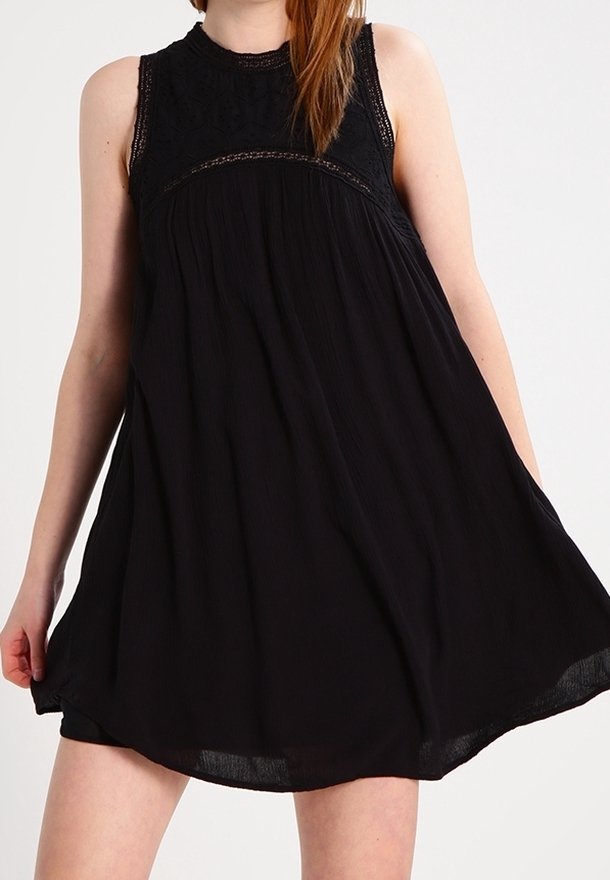 Femme portant une robe noire sans manches avec des détails en dentelle sur le haut, tenant légèrement la jupe fluide vers l'extérieur.