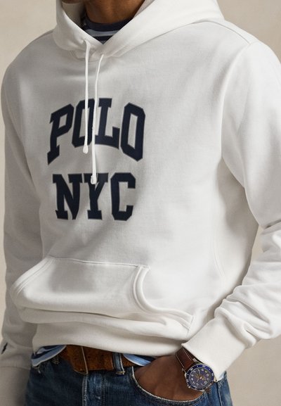 Άντρας που φοράει λευκό φούτερ με την επιγραφή «POLO NYC», μπλε τζιν, καφέ ζώνη και ρολόι χειρός με μπλε καντράν και καφέ δερμάτινο λουράκι.