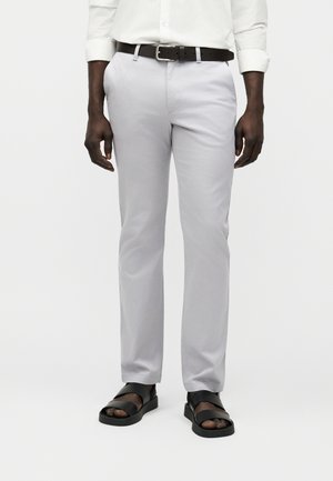 Homme portant un pantalon gris clair, une ceinture en cuir noire, une chemise blanche avec manches retroussées et des sandales ouvertes noires, debout devant un fond blanc.