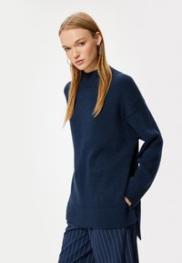Maglione a maglia blu navy con colletto alto, spalle scese, spacchi laterali e polsini e orlo a coste. Abbinato a pantaloni con righe sottili.
