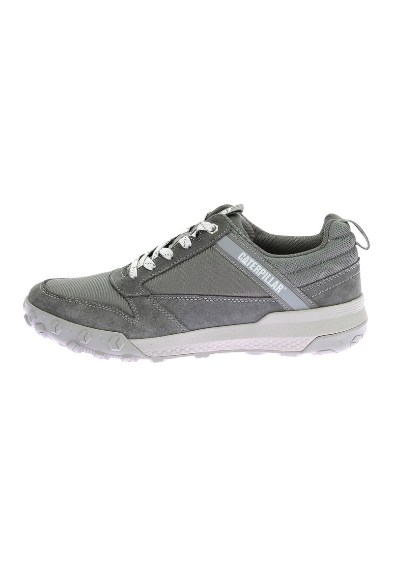 Zapatilla deportiva gris con una mezcla de materiales de malla y ante, suela de goma texturizada, diseño con cordones y un logotipo prominente en el costado.
