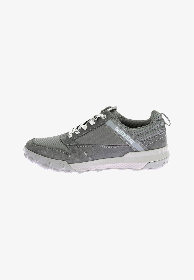 Zapatilla deportiva gris con una mezcla de materiales de malla y ante, suela de goma texturizada, diseño con cordones y un logotipo prominente en el costado.